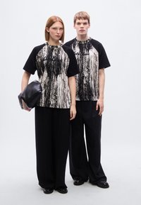 Ασπρόμαυρα γραφικά t-shirts με αφηρημένα σχέδια, σε συνδυασμό με μαύρο παντελόνι με φαρδιά μπατζάκια και αξεσουάρ, πάνω σε ανοιχτό γκρι φόντο.