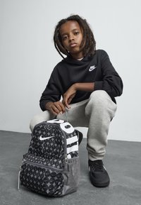 Νέος με κοτσιδάκια κάθεται σε γκρι πάτωμα, φορώντας ένα μαύρο hoodie της Nike, γκρίζα φόρμα, μαύρα αθλητικά παπούτσια και κρατώντας ένα σχέδιο σακίδιο της Nike.