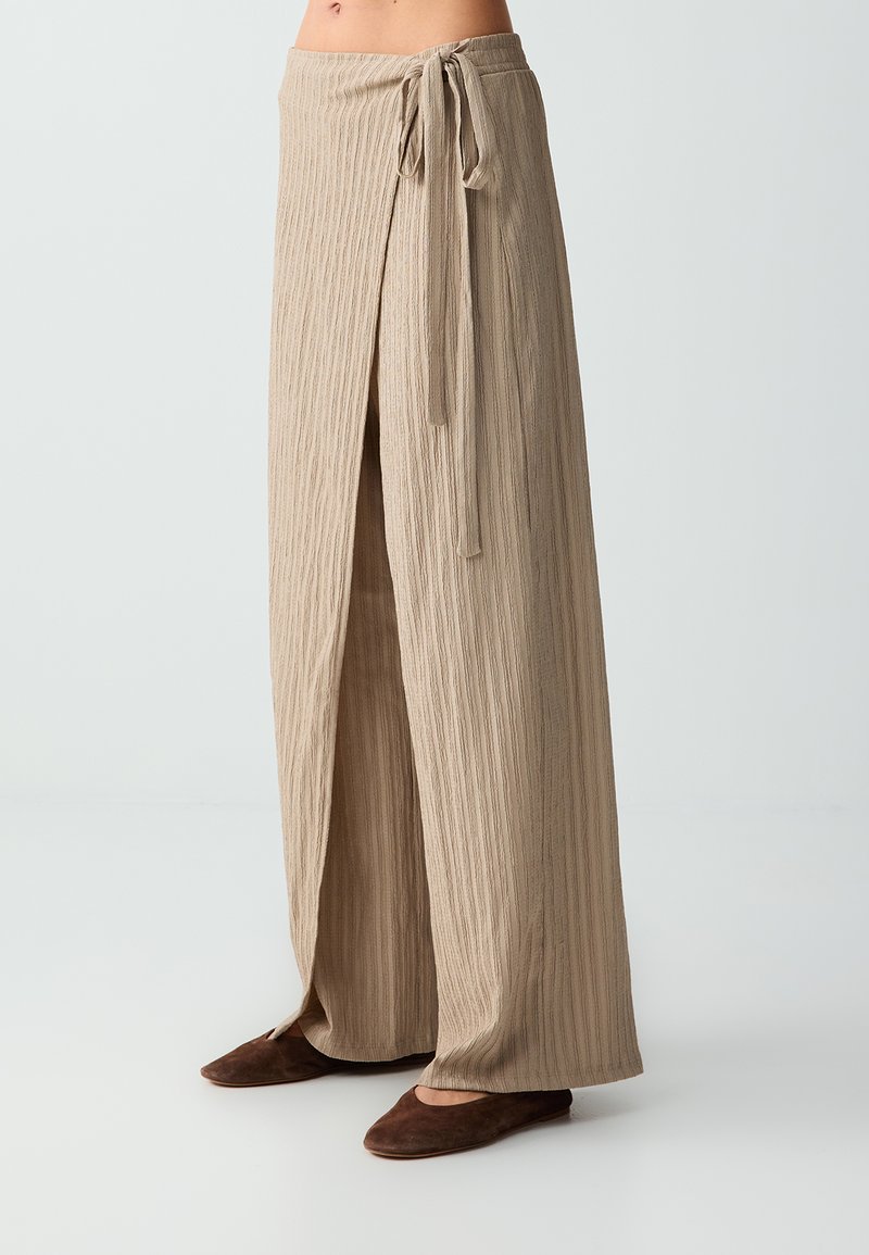 Pantalon beige à jambes larges texturé avec fente frontale et taille à nouer sur le côté, porté avec des chaussures plates marron foncé, visible de la taille aux pieds.
