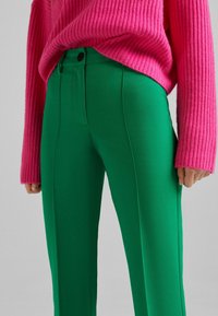 Gröna skräddarsydda byxor med sidosömmar och tvåfärgade svarta knappar, tillsammans med en ribbad rosa tröja. Slätt, elastiskt tyg.