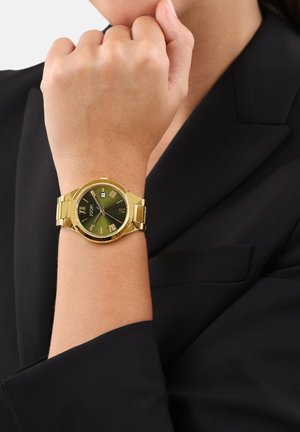 Person trägt eine goldene Uhr mit grünem Ziffernblatt und römischen Ziffern an ihrem linken Handgelenk, gekleidet in einem schwarzen Blazer.
