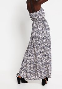 Kvinna i en ärmlös, mönstrad maxiklänning med svarta klacksandaler, stående med armarna lyfta mot en vit bakgrund.