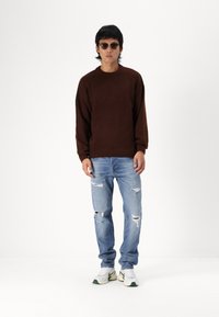 Brun strikket sweater, lyseblå ripped jeans og hvide sneakers med grønne accenter. Modellen bærer solbriller og står foran en ensfarvet baggrund.