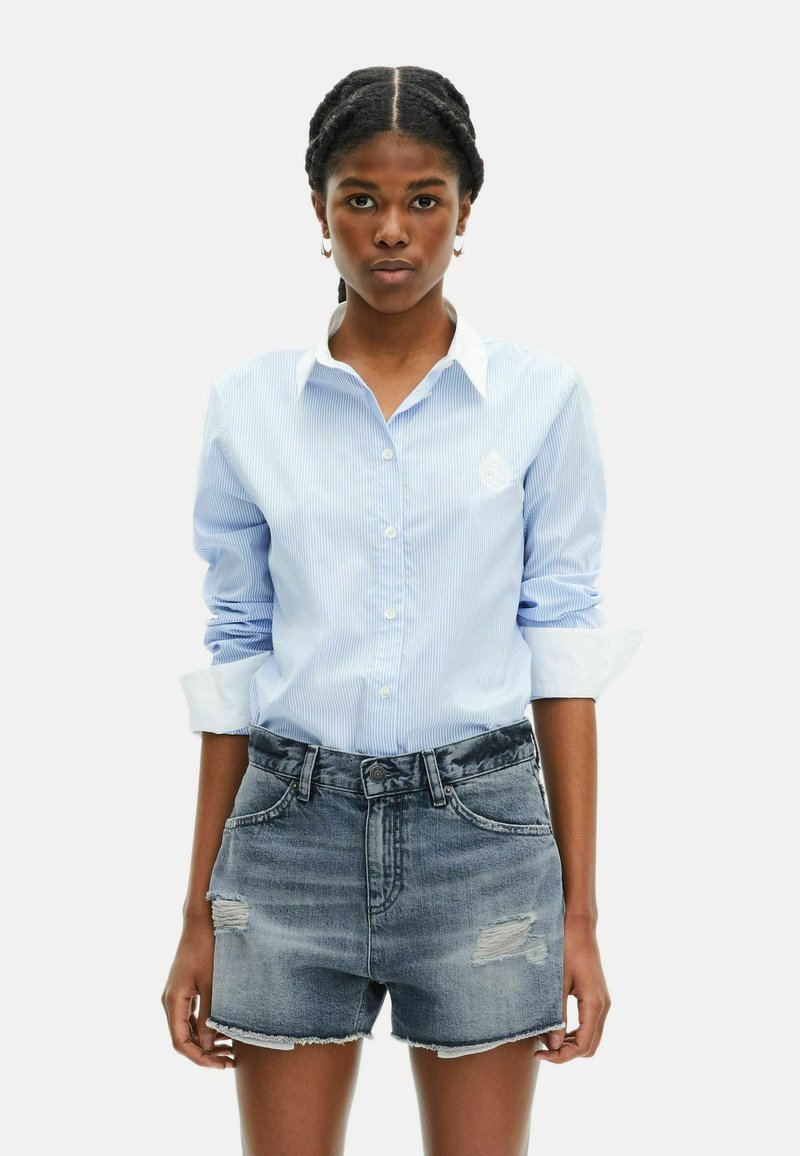 Jeune femme avec des cheveux tressés portant une chemise à boutons rayée bleu clair et un short en denim déchiré, se tenant devant un fond blanc.