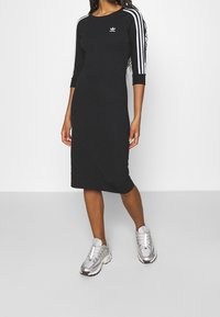 Mujer vestida con un vestido midi negro de Adidas con rayas blancas en las mangas y zapatillas plateadas de Adidas, de pie contra un fondo liso.