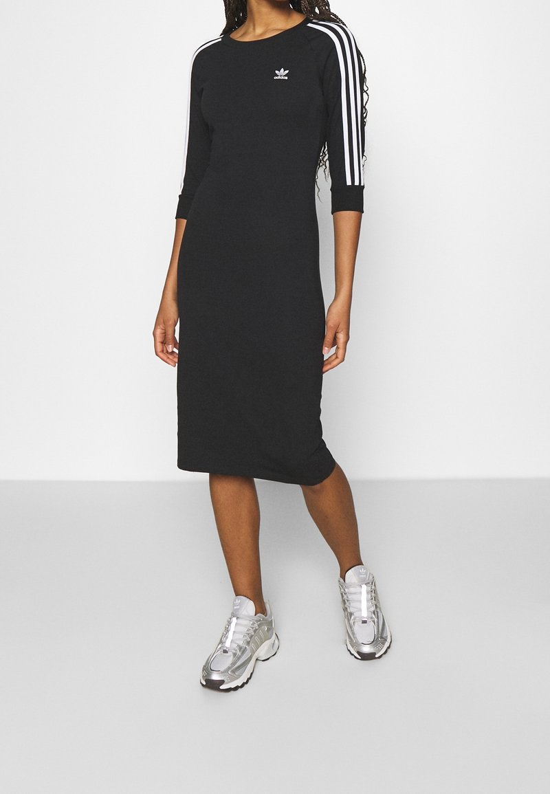 Mujer vestida con un vestido midi negro de Adidas con rayas blancas en las mangas y zapatillas plateadas de Adidas, de pie contra un fondo liso.