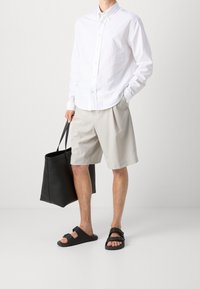 Camicia bianca con bottoni e logo ricamato, pantaloni corti beige, sandali piatti neri e una borsa a tracolla nera. Tessuti lisci, design informale.
