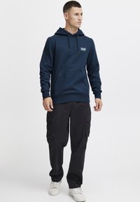 Felpa blu navy con tasca frontale e cappuccio regolabile, abbinata a pantaloni cargo neri e sneakers bianche. Design semplice e casual.