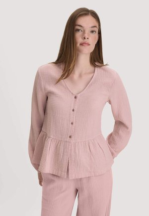 Jeune femme aux longs cheveux bruns porte un chemisier rose clair texturé à boutons avec basque et pantalon assorti sur fond uni.