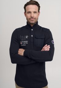Tengerészkék kötött pulóver magas gallérral, gombos nyakkal és mellzsebbel. Hímzett szöveggel és logókkal díszített. Bordás mandzsettákkal és aljjal rendelkezik.