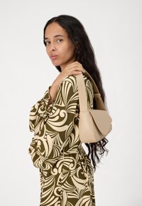 Geanta de piele bej cu un design curbat, mânere subțiri și închidere cu clapetă. Modelul poartă o rochie imprimată cu un model verde și crem.