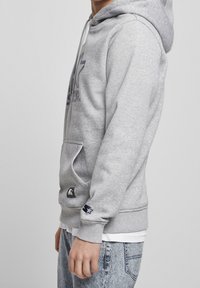 Grijze sweatshirt met capuchon, met een groot logo op de voorkant, een voorzak en geribde manchetten. Draagt over een wit shirt met spijkerbroek.