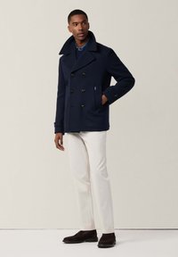 Abrigo de lana marinero en color azul marino con cierre de doble botonadura, gran cuello y botones negros; combinado con pantalones blancos y zapatos marrones.