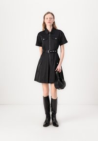Vestido preto de mangas curtas com fecho éclair na frente, gola de camisa, dois bolsos no peito, cintura com cinto, saia plissada, combinado com botas pretas até ao joelho e uma pequena mala.