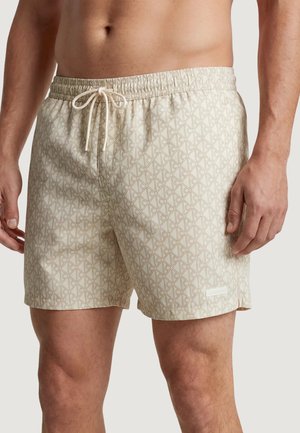Buste et jambes masculins portant un short de bain beige à motifs avec une ceinture élastique et un cordon à l'avant, debout devant un fond neutre.