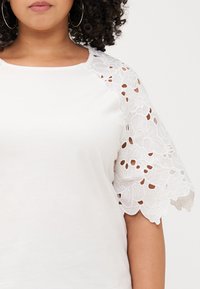 Close-up of a person wearing a white shirt with lace-trimmed sleeves, featuring curly hair and hoop earrings against a plain background.