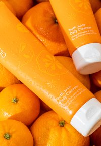 Deux tubes orange de "Truth Juice Daily Cleanser" sont partiellement immergés parmi des oranges fraîches, arborant une finition mate et des bouchons blancs.
