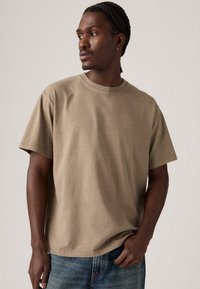 Mann med flettehår iført en beige Levi's t-skjorte og blå jeans, ser til siden mot en lys bakgrunn.