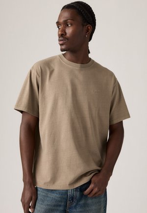 Hombre con pelo trenzado que lleva una camiseta beige de Levi's y jeans azules, mirando hacia un lado contra un fondo claro y uniforme.