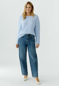 Frau steht mit einem hellblauen Pullover, hochtaillierten Jeans mit weitem Bein und weißen spitzen Absatzschuhen vor einem schlichten Hintergrund.