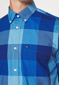 Primer plano de una persona que lleva una camisa de cuadros azul, con botones, cuello y un bolsillo en el pecho que presenta un pequeño logotipo de bandera.