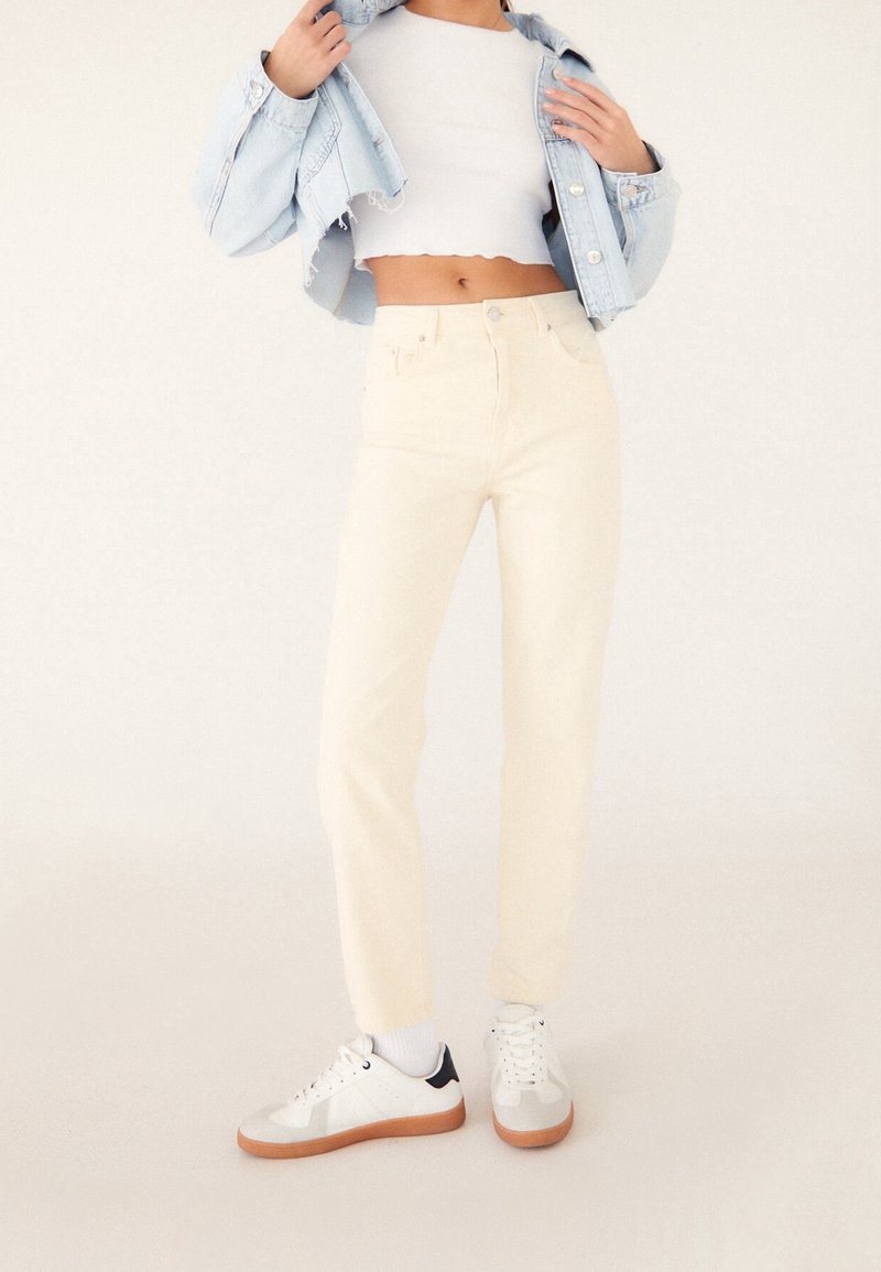 Femme portant un pantalon crème taille haute, un crop top blanc, une veste en jean bleue claire courte, des chaussettes blanches et des baskets blanches avec une semelle en gomme.