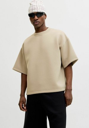 Mężczyzna ubraną w beżową, oversize koszulę z krótkim rękawem, czarne spodnie, wzorzysty biały czapka, czarne okulary przeciwsłoneczne oraz małą kolczyk w kształcie kółka na jednolitym tle.