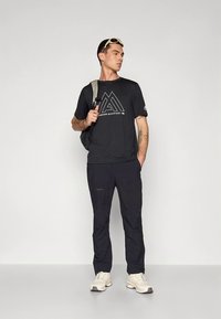 Must lühikeste varrukatega t-särk valge graafilise logoga, kombineeritud tumedate pükstega. Kandmas kerget seljakotti ja beeže spordijalatseid.