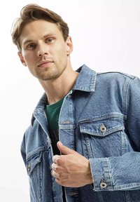 Jasnoniebieska kurtka dżinsowa z klasycznym krojem, kieszeniami na piersi i zapięciem na guziki. Noszona na zielonym t-shircie, o gładkiej, lekko wyblakłej fakturze.