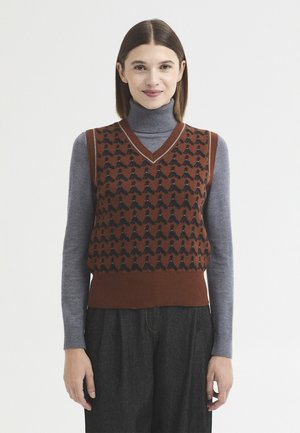 Femme aux cheveux bruns et lisses porte un gilet en laine marron à motifs par-dessus un col roulé gris et un pantalon gris foncé, debout devant un fond blanc.