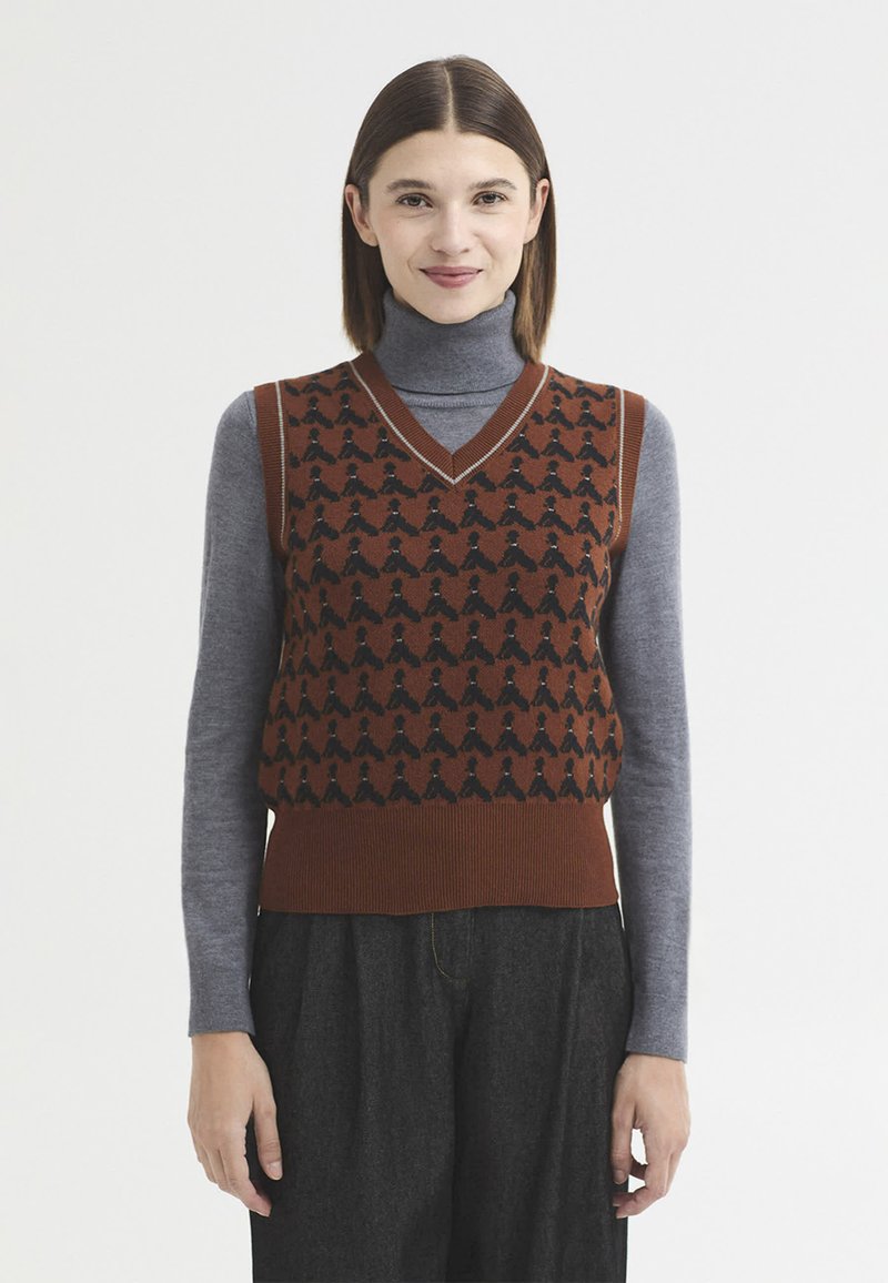 Femme aux cheveux bruns et lisses porte un gilet en laine marron à motifs par-dessus un col roulé gris et un pantalon gris foncé, debout devant un fond blanc.