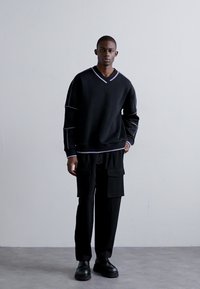 Maglione nero a V con rifiniture a contrasto, abbinato a pantaloni cargo neri con tasche. Il modello è in piedi su uno sfondo neutro, indossando scarpe nere.