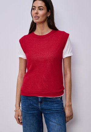 Kobieta ubrana w bezrękawnik z czerwonego dzianego swetra na białej koszulce z krótkim rękawem i niebieskich dżinsach, stojąca na jednolitym tle.