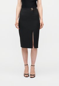 Γυναίκα που φοράει μαύρη pencil φούστα μήκους μέχρι το γόνατο με ανοίγμα στη μπροστινή πλευρά, λεπτομέρεια με χρυσή αγκράφα στη μέση, μαύρο αμάνικο τοπ και μαύρα πέδιλα με λουράκια.