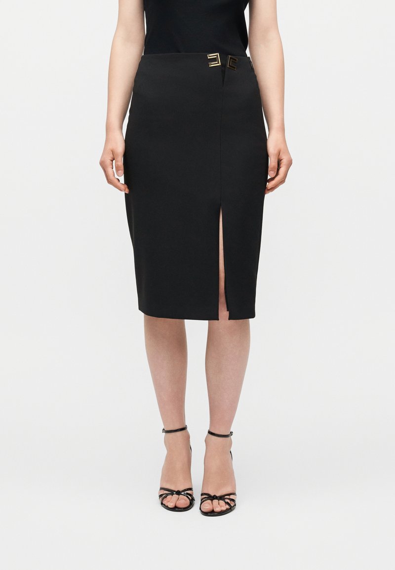 Γυναίκα που φοράει μαύρη pencil φούστα μήκους μέχρι το γόνατο με ανοίγμα στη μπροστινή πλευρά, λεπτομέρεια με χρυσή αγκράφα στη μέση, μαύρο αμάνικο τοπ και μαύρα πέδιλα με λουράκια.