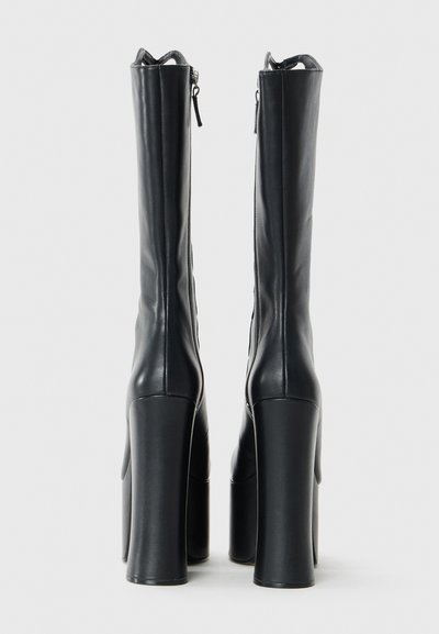 Bottes noires à plateforme hauteur genou avec des talons épais et des fermetures éclair latérales, se tenant debout sur un fond blanc uni.