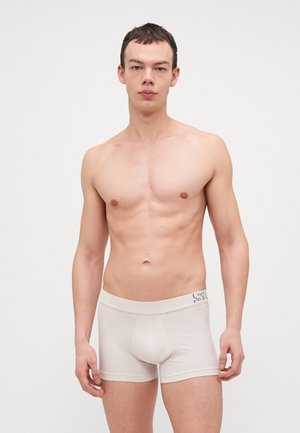 Jeune homme debout devant un fond uni, portant un boxer beige clair, regardant devant avec une expression neutre.