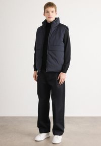 Gilet imbottito blu con colletto alto e due tasche frontali, indossato sopra un maglione nero, abbinato a pantaloni neri e sneakers bianche.