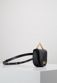 Petit sac bandoulière en cuir noir avec devant matelassé, anse chaîne dorée, sangle ajustable et logo décoratif doré sur une surface blanche.