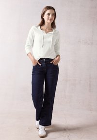 Witte lange mouwen shirt met een capuchon, donkerblauwe wijde jeans en witte sneakers. Model poseert met handen in de zakken tegen een neutrale achtergrond.