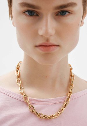 Collar de cadena dorada con grandes eslabones entrelazados. Lleva puesto sobre una camiseta rosa claro con un escote amplio, que muestra una textura lisa y brillante.