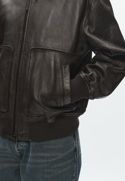 Sötétbarna bőrdzseki bordázott mandzsettákkal és szegéllyel, elöl cipzárral és nagy oldalzsebekkel, kék farmernadrág fölött viselve.