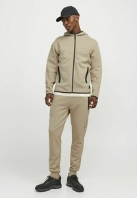 Bežová mikina na zips s čiernymi akcentmi a bočnými vreckami, spárovaná s rovnakými teplákmi. Čierna čiapka a trekingové topánky dokončujú outfit.