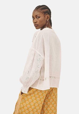 Blusa de lino blanco con mangas largas tipo globo que presentan bordado de ojal, combinada con pantalones de estampado floral mostaza. Diseño simple y relajado.