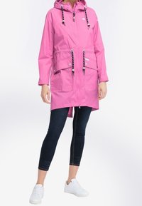 Kevyt vaaleanpunainen sadetakki hupulla, nauhakiinnityksillä, etutaskuilla ja vetoketjulla, päällä tummat legginsit ja valkoiset kengät.