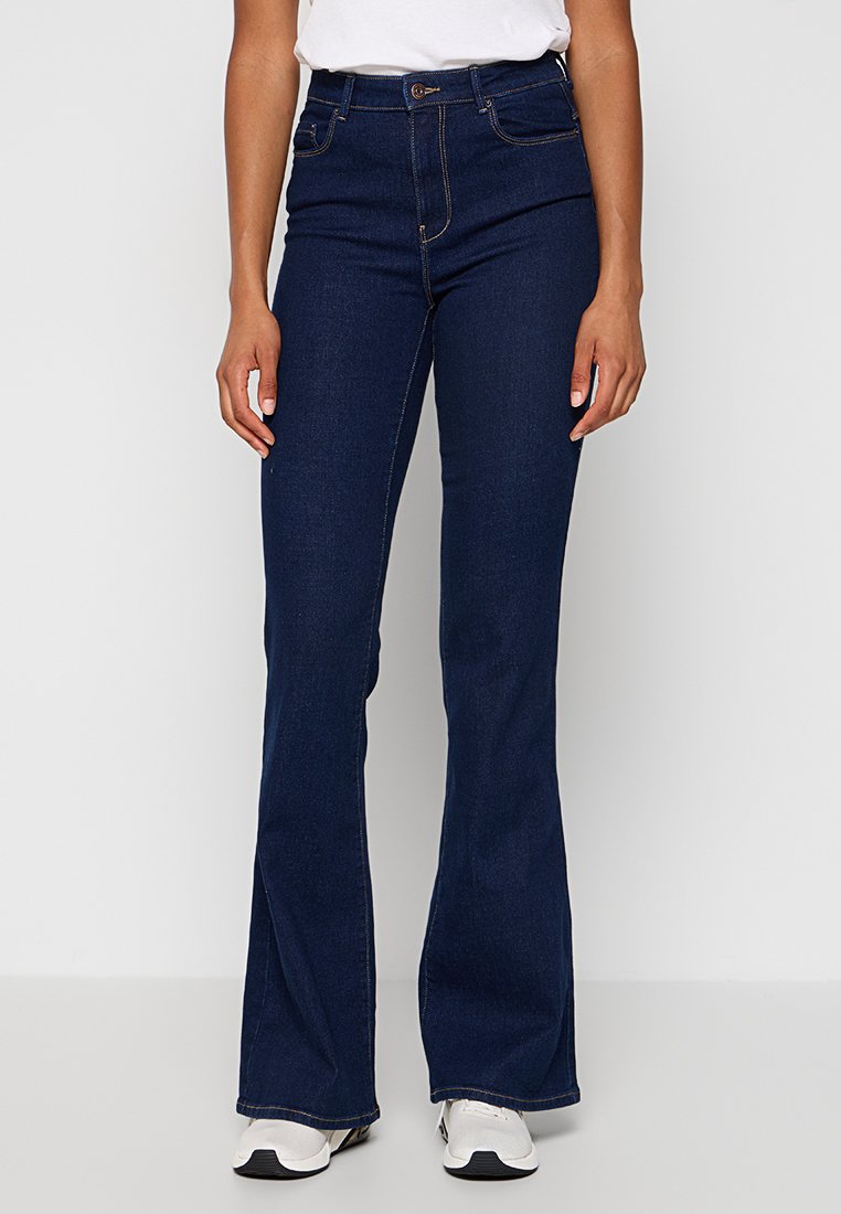 Jeans évasés taille haute bleu foncé avec poches avant, coutures contrastées et texture en denim lisse, associés à des baskets blanches.