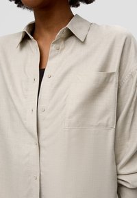 Beige button-up shirt made of textured fabric. Features a single chest pocket and standard collar. Buttons in a lighter color.