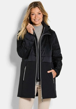 Lächelnde Frau mit blonden Haaren, die einen schwarzen, gepolsterten Mantel mit Reißverschlusstaschen über einem grauen Pullover und beigen Hosen trägt.