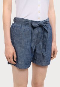 Blå denim shorts med ett knutet tygbälte, framfickor och uppvikta ben, bärs med en vit knäppt topp.