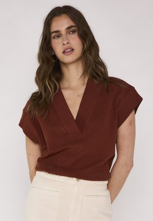 Giovane donna con capelli lunghi e ondulati, indossa una maglia a V color ruggine e pantaloni chiari a vita alta, guarda leggermente di lato su uno sfondo semplice.